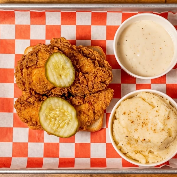Crispy fried chicken with pickles on white bread, mashed potatoes, and gravy on a metal tray.