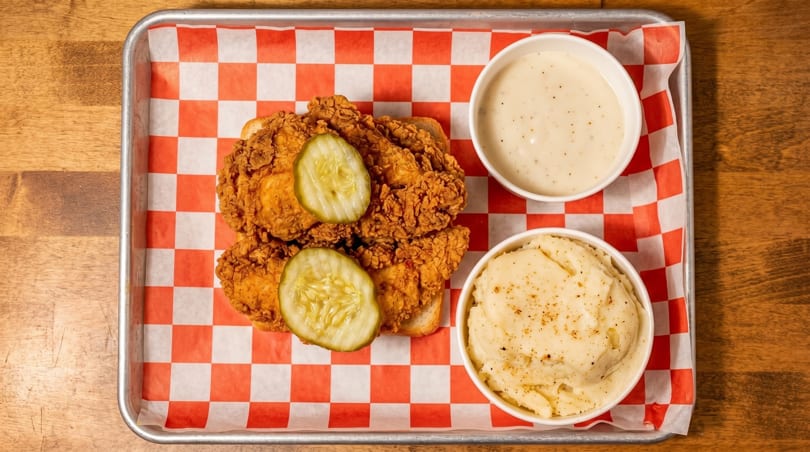 Crispy fried chicken with pickles on white bread, mashed potatoes, and gravy on a metal tray.