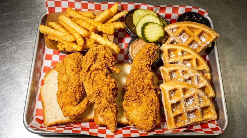 Crispy fried chicken tenders, crinkle-cut fries, waffles, pickles, and dipping sauces on a tray.