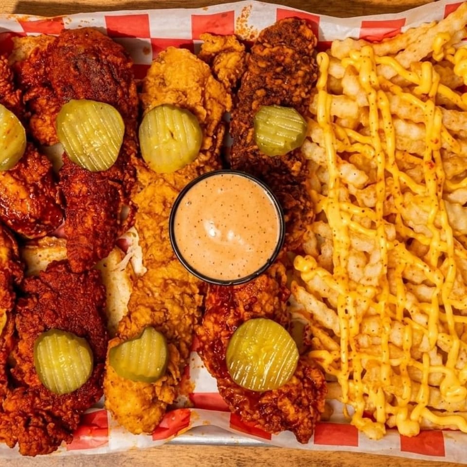 A metal tray with spicy chicken tenders, pickles, dipping sauce, and cheese fries.