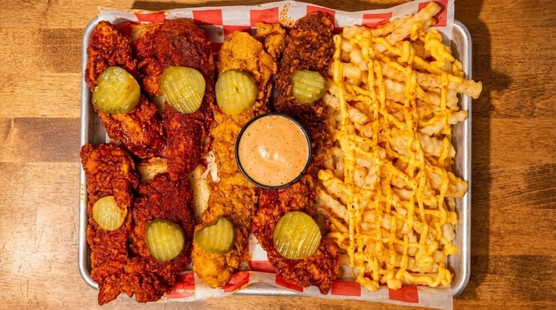 A metal tray with spicy chicken tenders, pickles, dipping sauce, and cheese fries.