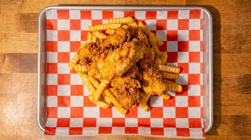 Crispy fried chicken tenders and crinkle-cut fries on red and white checkered paper in a metal tray.