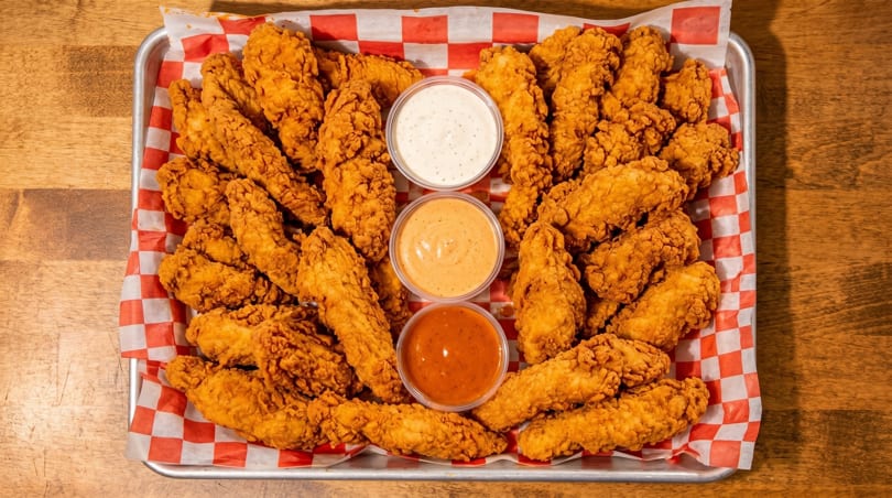 A metal tray filled with crispy fried chicken tenders and three dipping sauces on checkered paper.