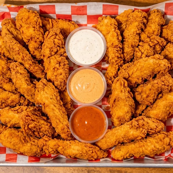 Crispy golden chicken tenders on a metal tray with red and white checkered paper, served with three dipping sauces.