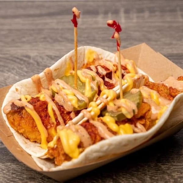 A crispy chicken taco with pickles, cheese sauce, and spicy mayo in a paper tray.