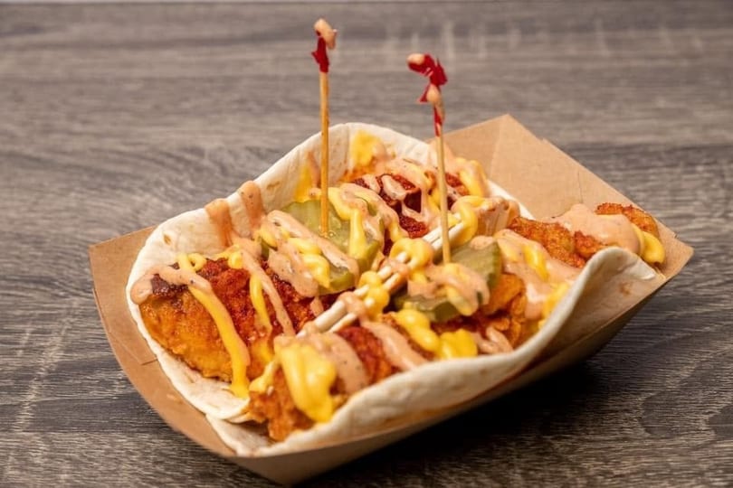 A crispy chicken taco with pickles, cheese sauce, and spicy mayo in a paper tray.